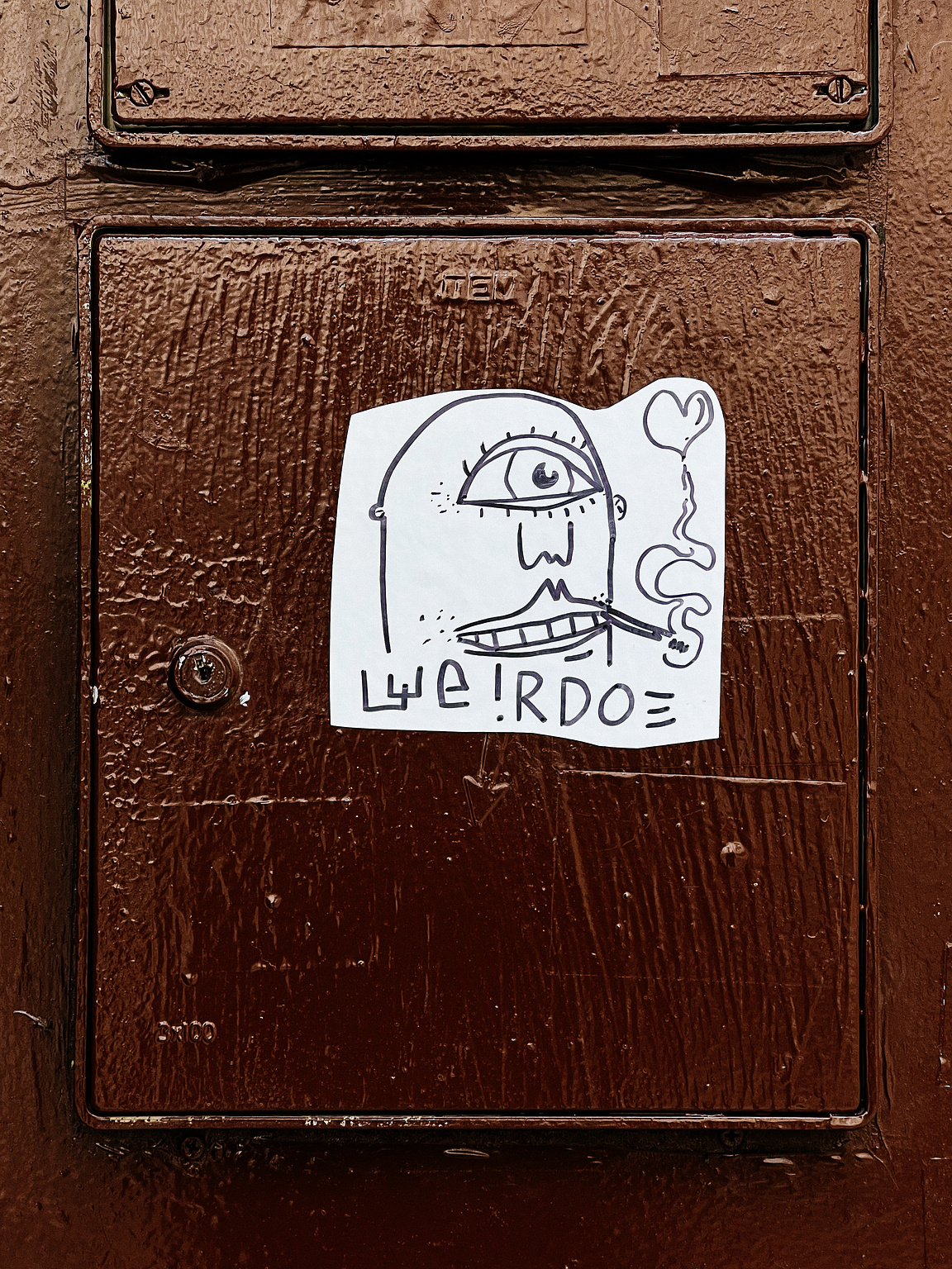A sticker with a doodle of a face and the word "WEIRDO" is affixed to a textured brown door.