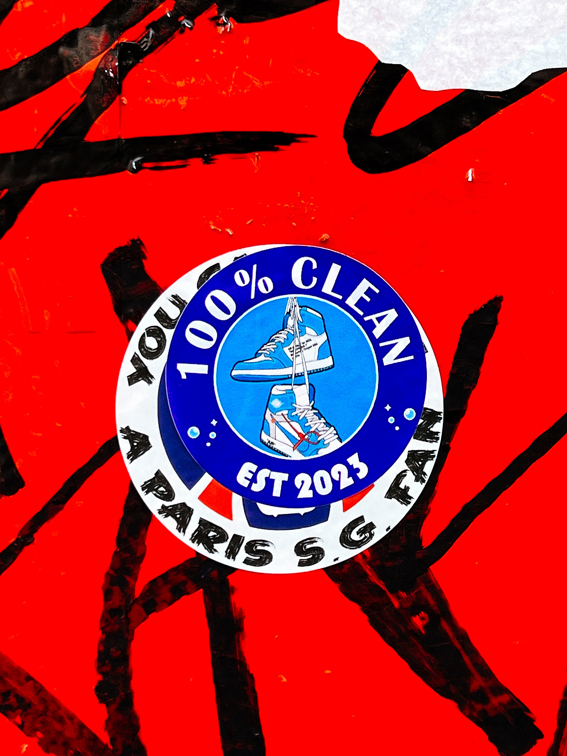A circular sticker on a red, textured background displays the text "100% CLEAN EST 2023" surrounding an image of a couple of sneakers.