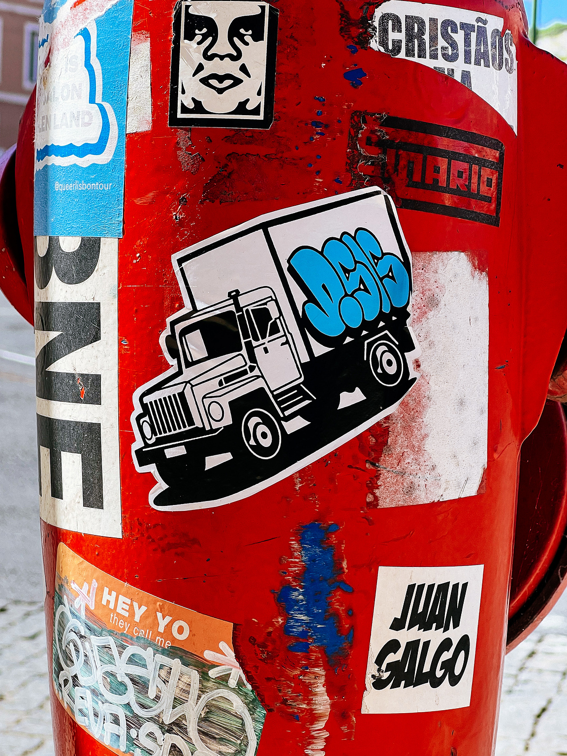 A red pole covered in various stickers, some depicting vehicles and faces, amidst urban surroundings.