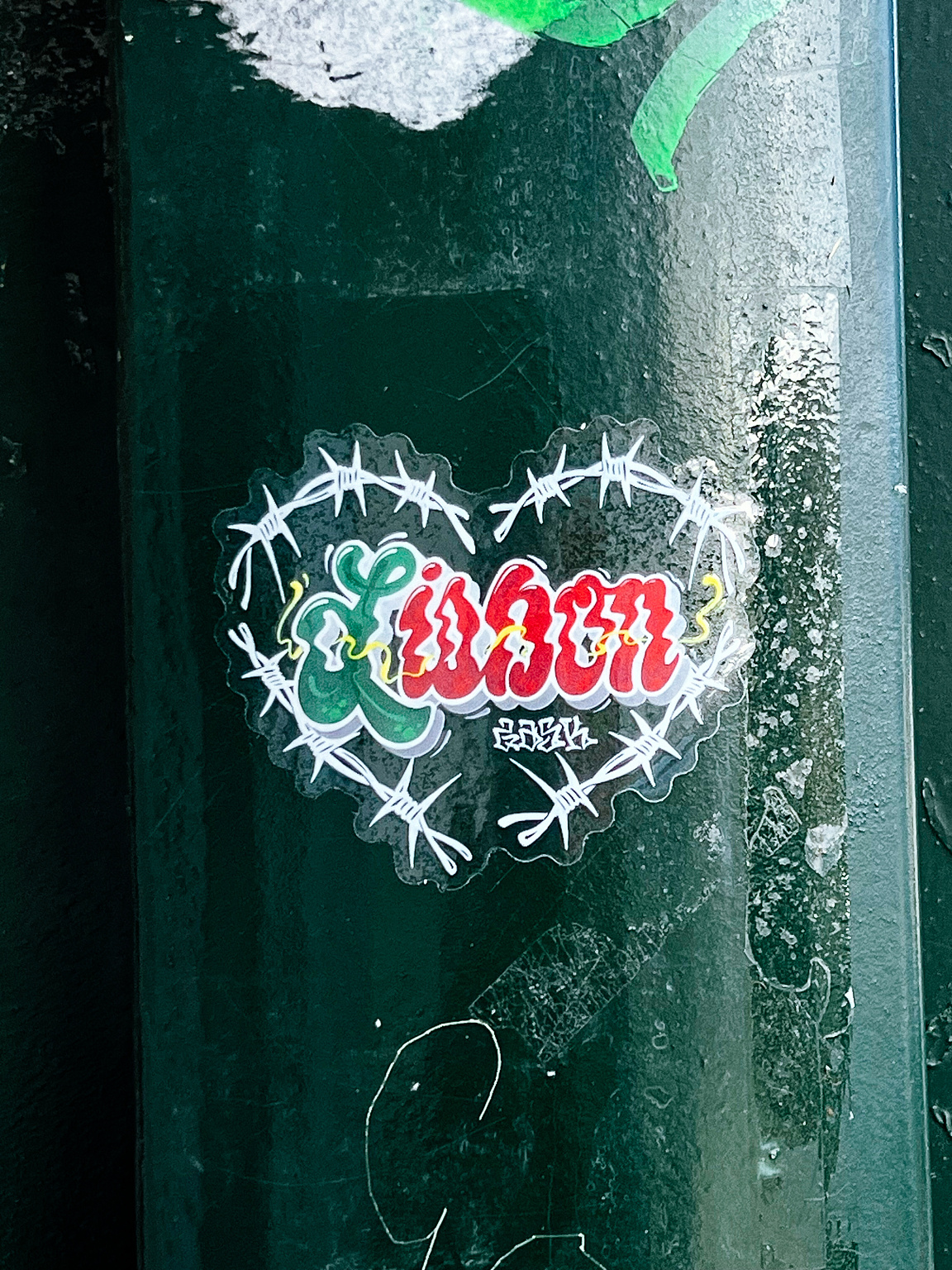A sticker depicting a stylized heart shaped by barbed wire, with "Lisbon" written in graffiti style, is adhered to a weathered metal surface.