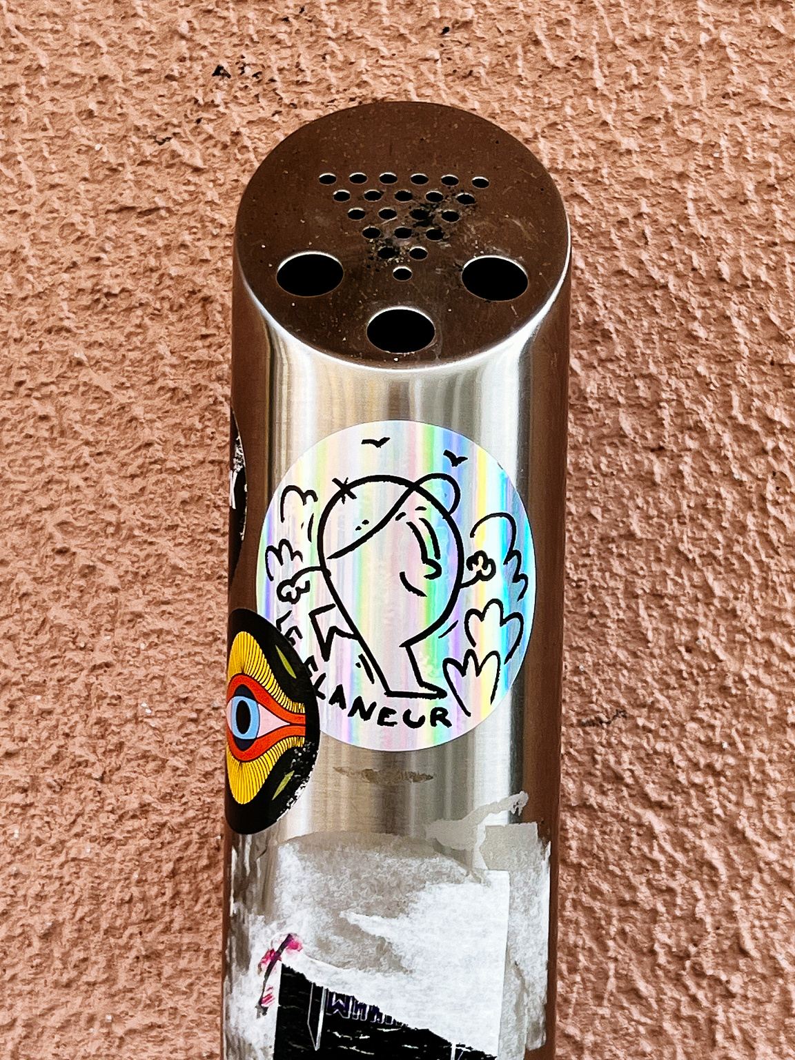 A cylindrical ashtray adorned with stickers and graffiti against a textured wall.