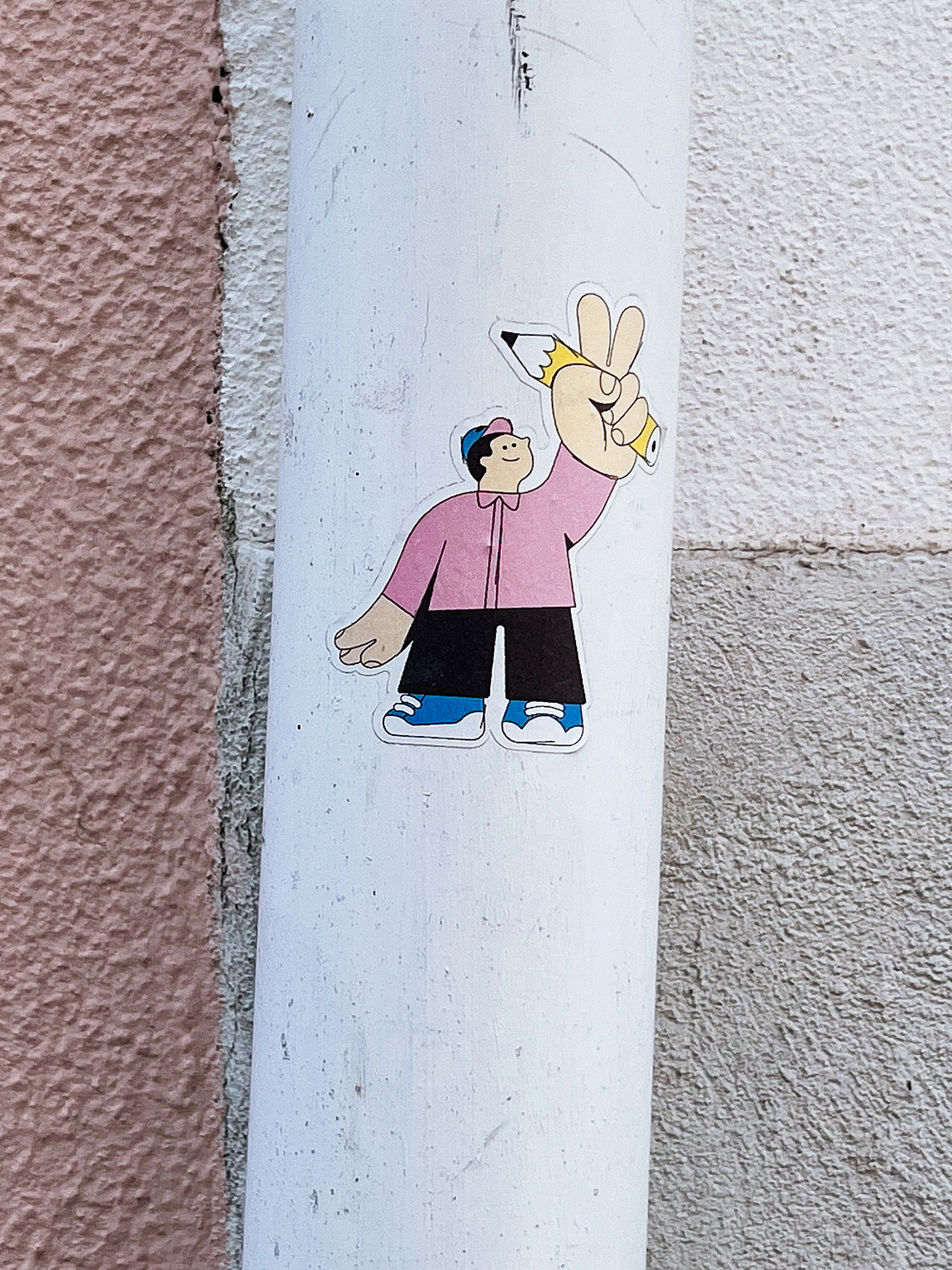 A sticker depicting a cartoon character making a peace sign, and holding a pencil, affixed to a white vertical street pipe against a pink wall.