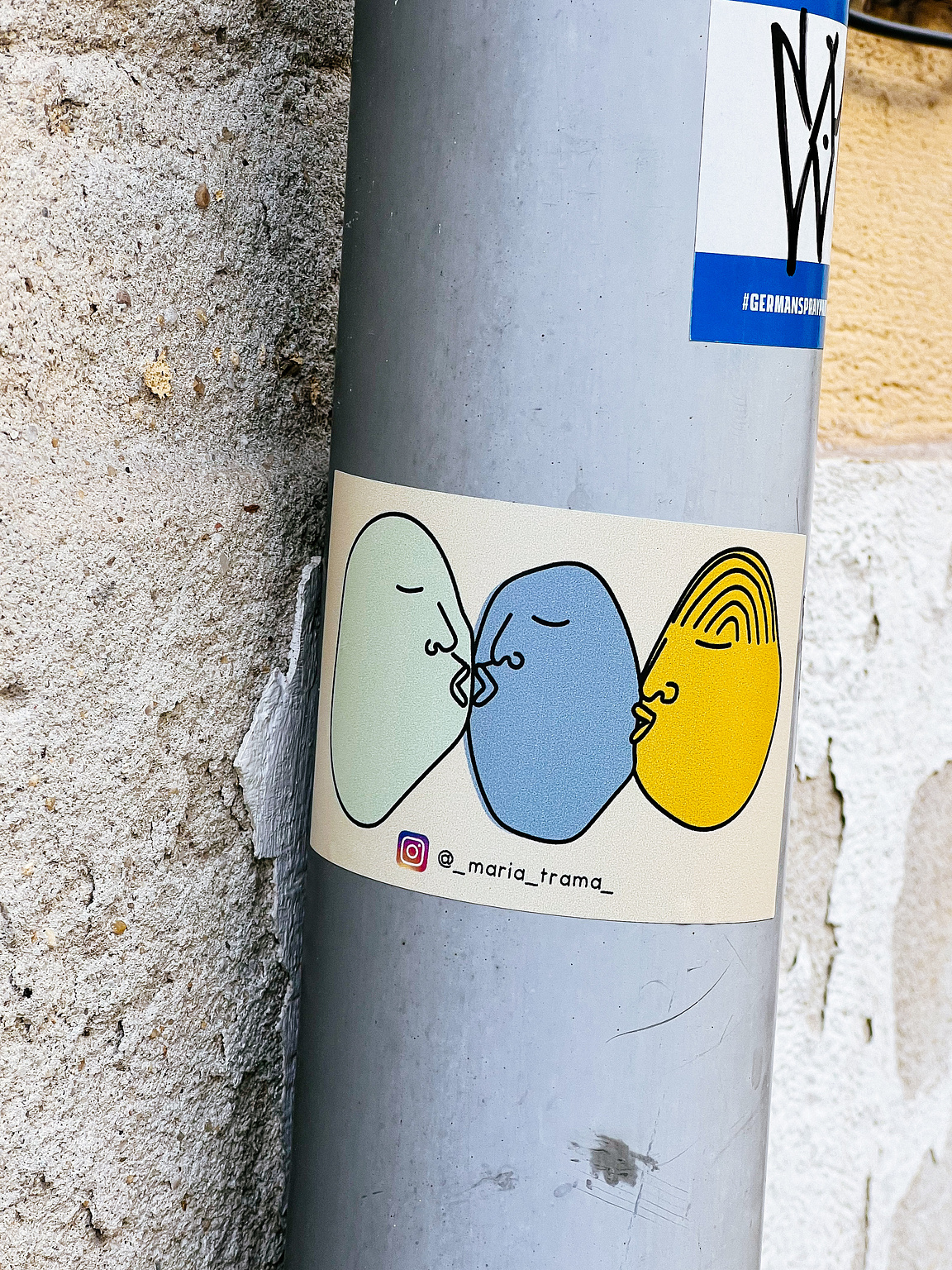 Three stylized faces in primary colors kiss each other on a sticker, affixed to a gray pole with rough texture. Instagram handle @_maria_trama_ is visible.