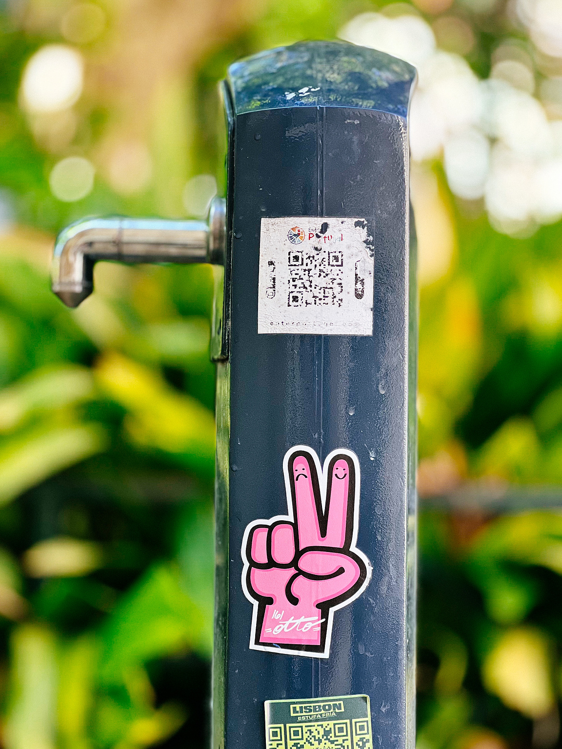 A metal pole with a tap, covered in faded stickers, against a blurred green foliage background.