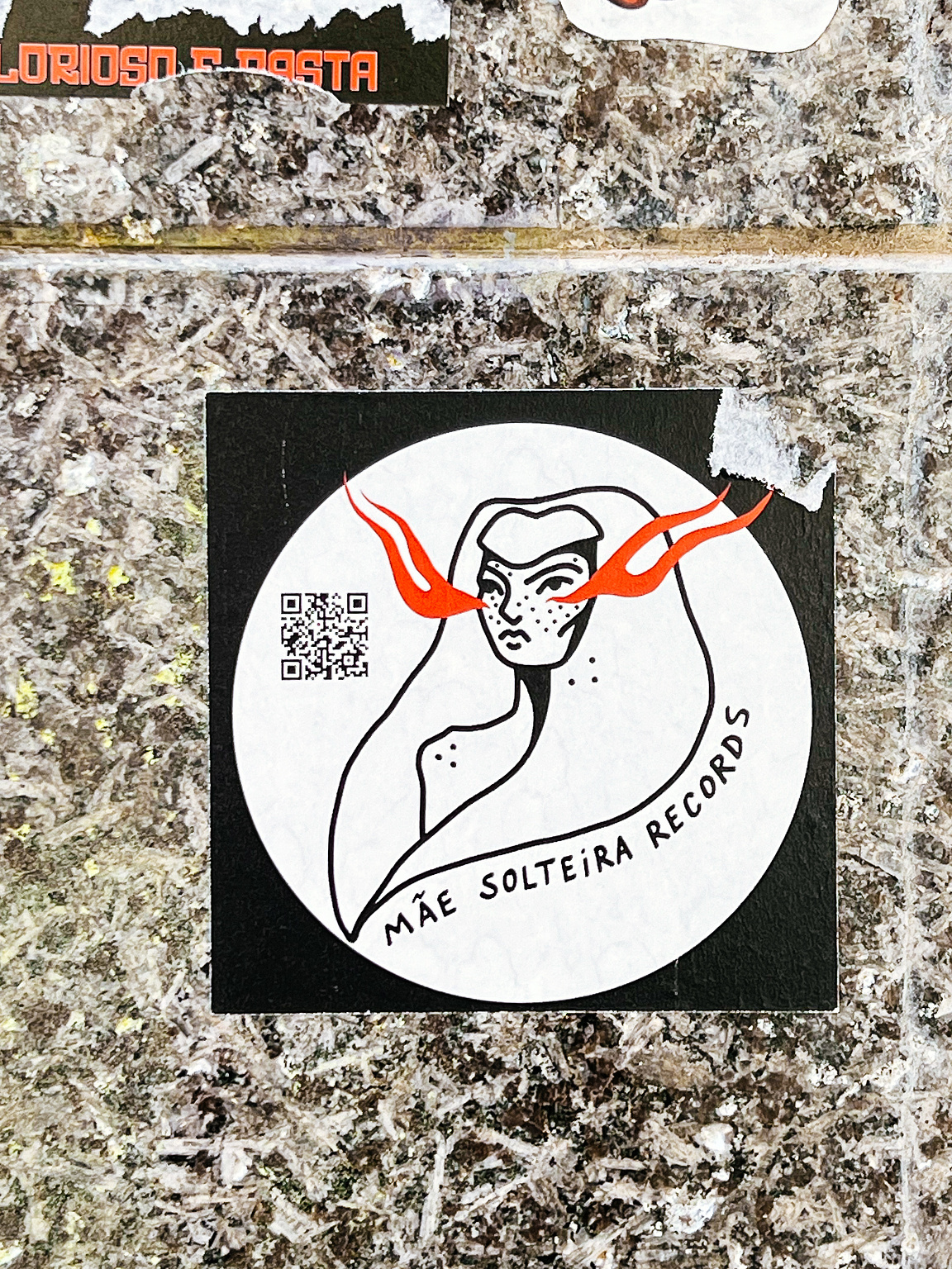 A sticker with a stylized female face and flowing hair, above the text "MÃE SOLTEIRA RECORDS," attached to a speckled wall.